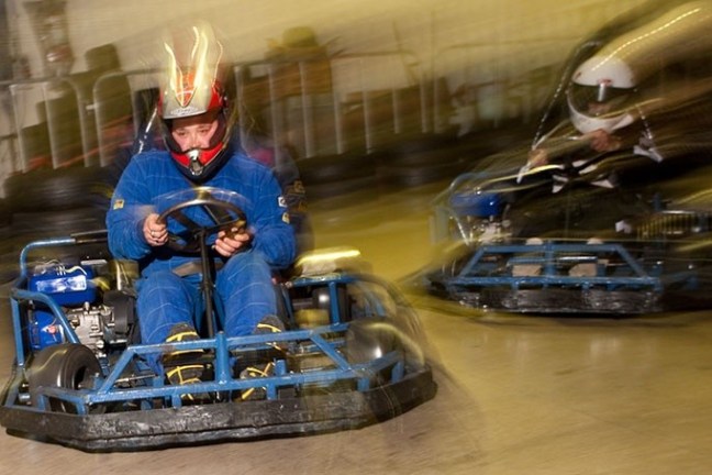 Two people racing go-karts indoors, wearing helmets and racing suits.
